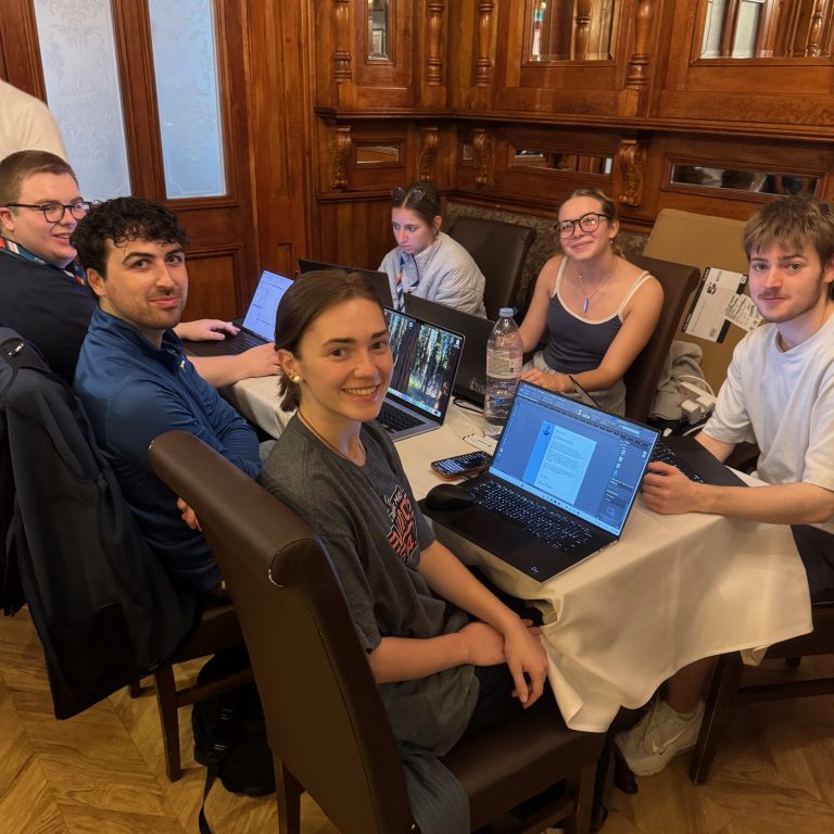Six people sit around a table with laptops and papers in a wood-paneled room, looking at the camera and working together.