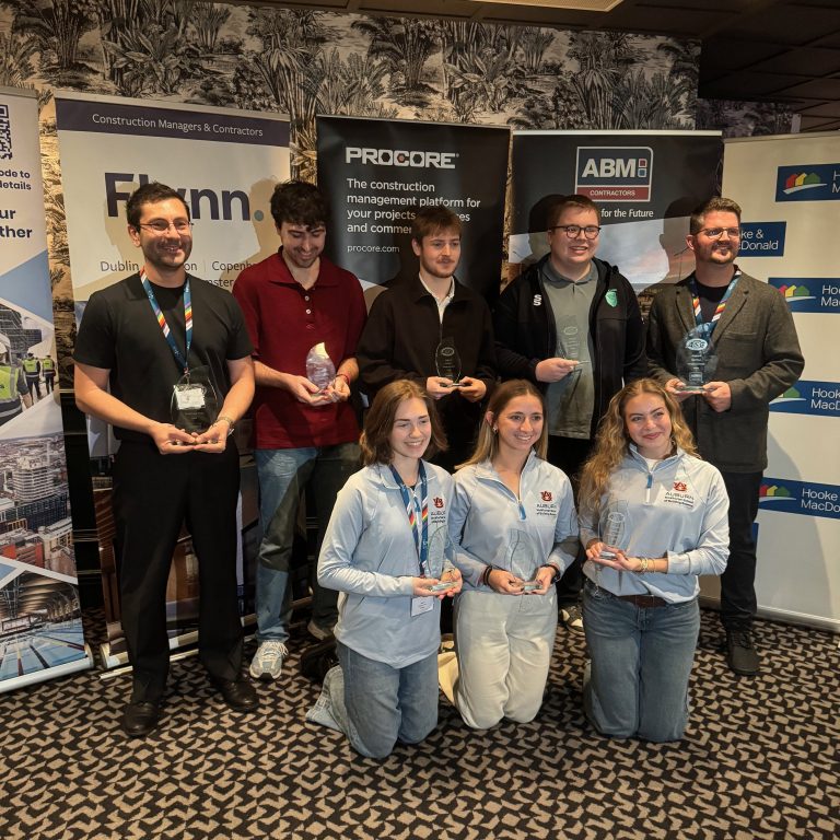A group of eight people, four standing and four kneeling, hold glass awards and pose in front of construction company banners at an indoor event.