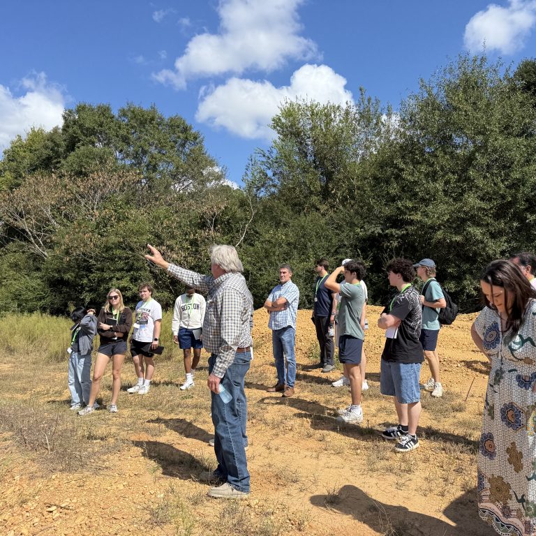 A group of people stands outdoors on a sunny day, listening to a man gesturing with his arm raised. Trees and blue sky with clouds are in the background.