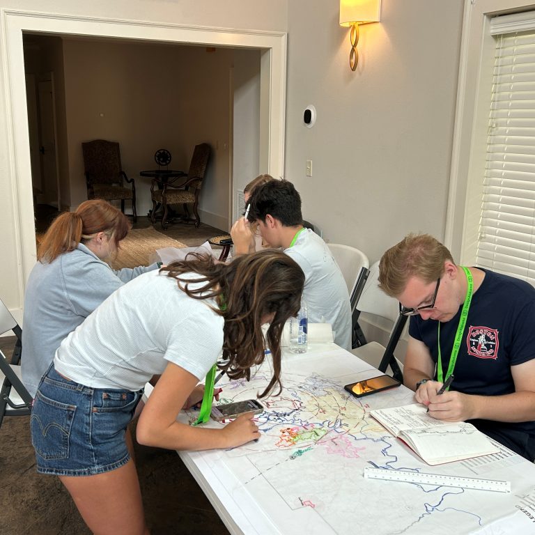 Four people work together at a table, examining maps and writing notes, while another person sits in the background of a well-lit room.