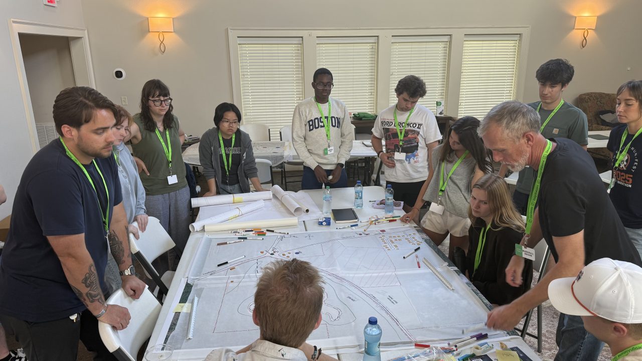 A group of people stands and sits around a large table covered with maps and papers, engaged in discussion in a well-lit room.