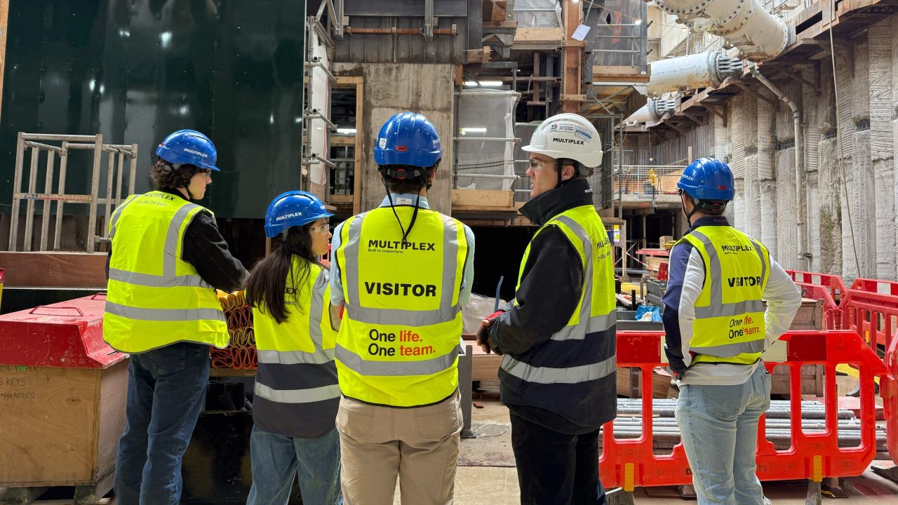 Five people in safety vests and helmets stand at a construction site, talking and observing the area; two vests are labeled “VISITOR.”.