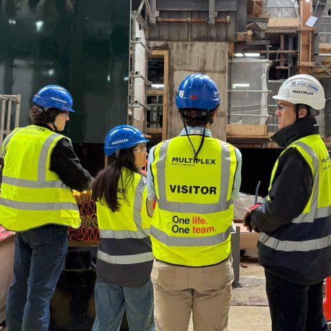 Five people in safety vests and helmets stand at a construction site, talking and observing the area; two vests are labeled “VISITOR.”.