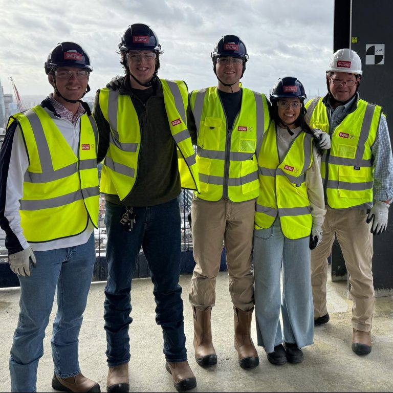 Five people wearing safety vests, helmets, gloves, and boots stand on a high-rise construction site with a city skyline in the background.