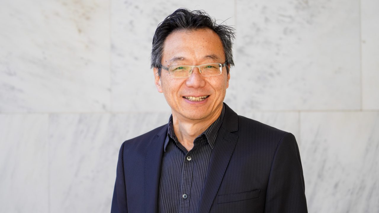 A man in a dark suit and striped shirt, wearing glasses, stands in front of a white marble wall, smiling at the camera.