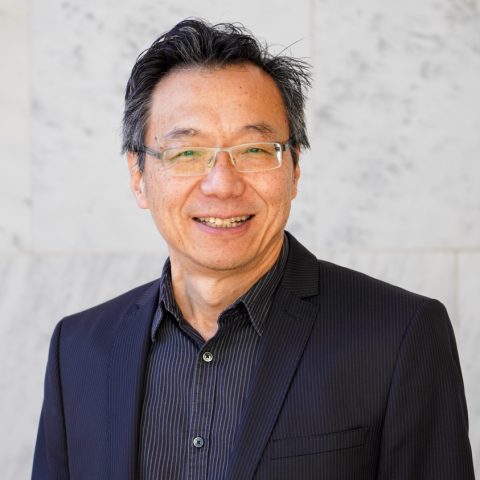 A man in a dark suit and striped shirt, wearing glasses, stands in front of a white marble wall, smiling at the camera.