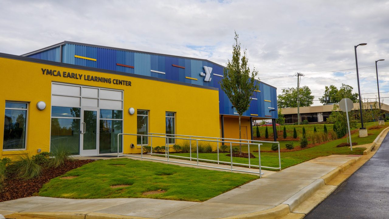 Exterior view of the YMCA Early Learning Center building with a yellow facade, glass entrance doors, and a wheelchair-accessible ramp on a cloudy day.