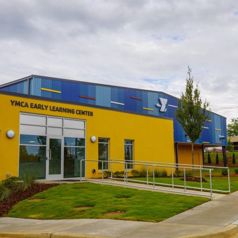 Exterior view of the YMCA Early Learning Center building with a yellow facade, glass entrance doors, and a wheelchair-accessible ramp on a cloudy day.