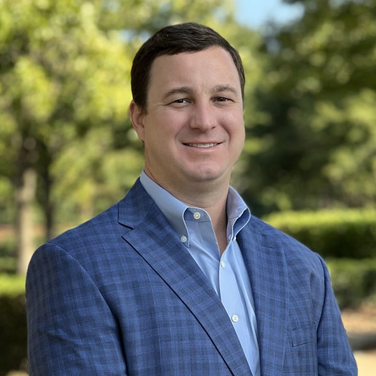 A man in a blue checked suit jacket stands outdoors in front of green trees and bushes, smiling at the camera.