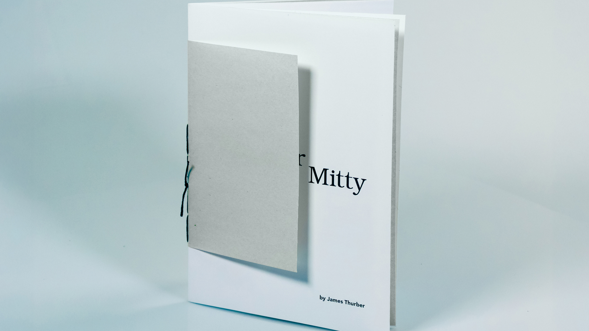 A booklet with a plain gray cover partially obscuring the title Mitty and the authors name James Thurber on a white page behind it, standing upright on a light surface.