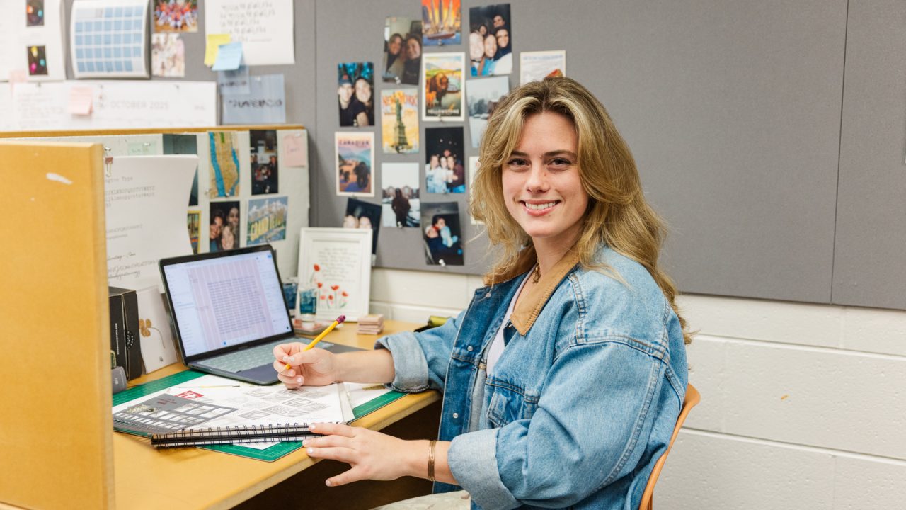 A young woman sits at a desk in a workspace, smiling at the camera while writing in a notebook; a laptop and various papers are on the desk.