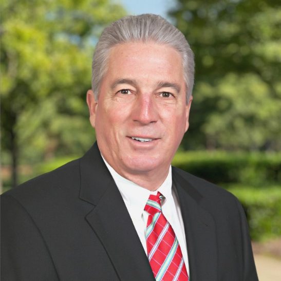 A middle-aged man with gray hair wearing a black suit, white shirt, and red striped tie, standing outdoors with trees and greenery in the background.