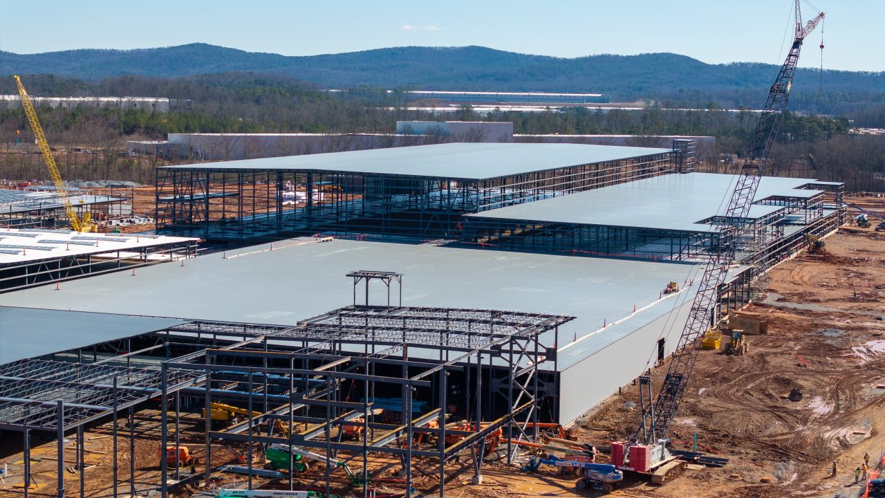 A large industrial building under construction with steel framework, cranes, and construction equipment on a dirt site, set against a backdrop of wooded hills.
