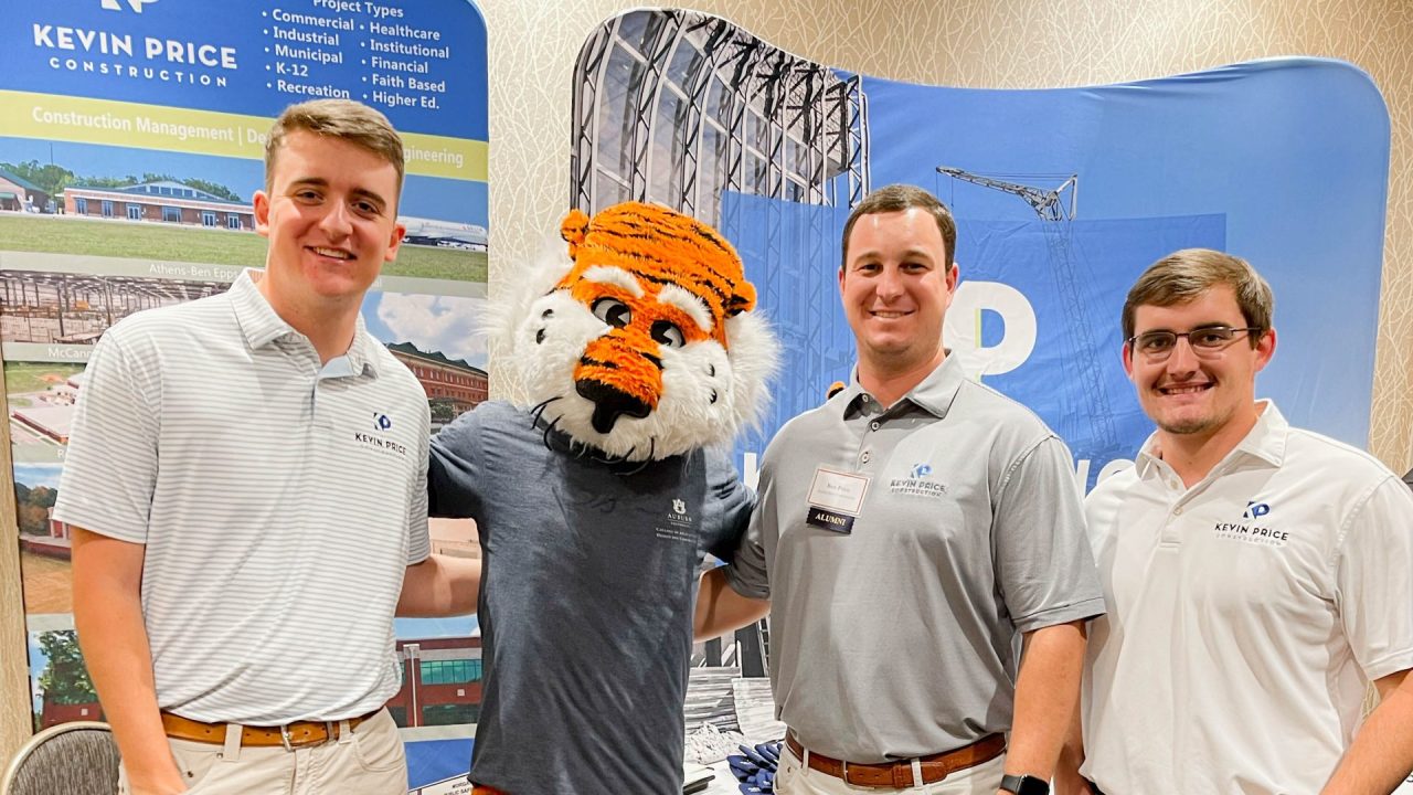 Three men in light-colored shirts and khaki pants stand next to a person in a tiger mascot costume in front of a Kevin Price Construction booth.