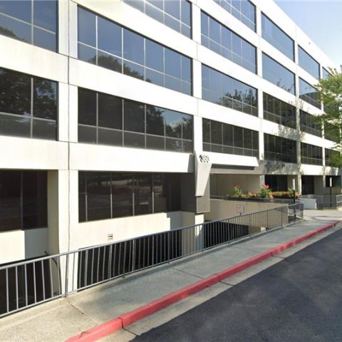 Four-story office building with large glass windows, a white and black exterior, and a ramp with railings along the front entrance. Trees are visible in the background.