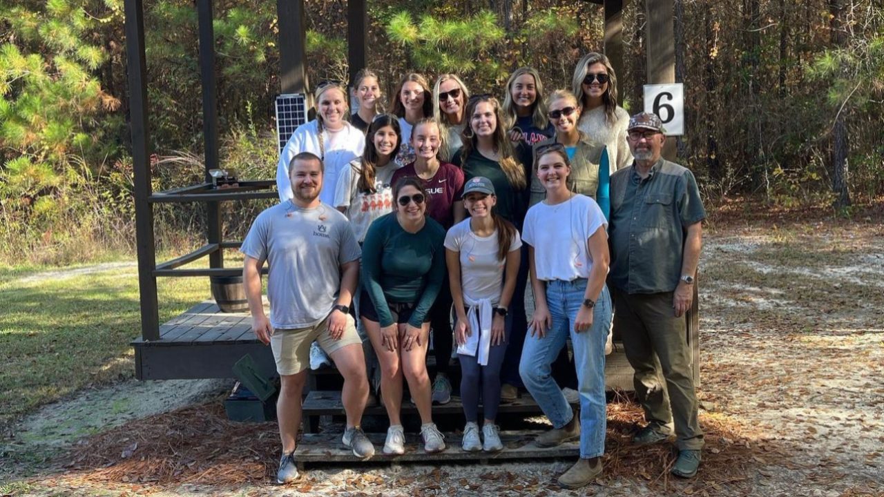 A group of 15 people pose for a photo in front of a wooden shelter labeled with the number 6, surrounded by trees and sunlight.