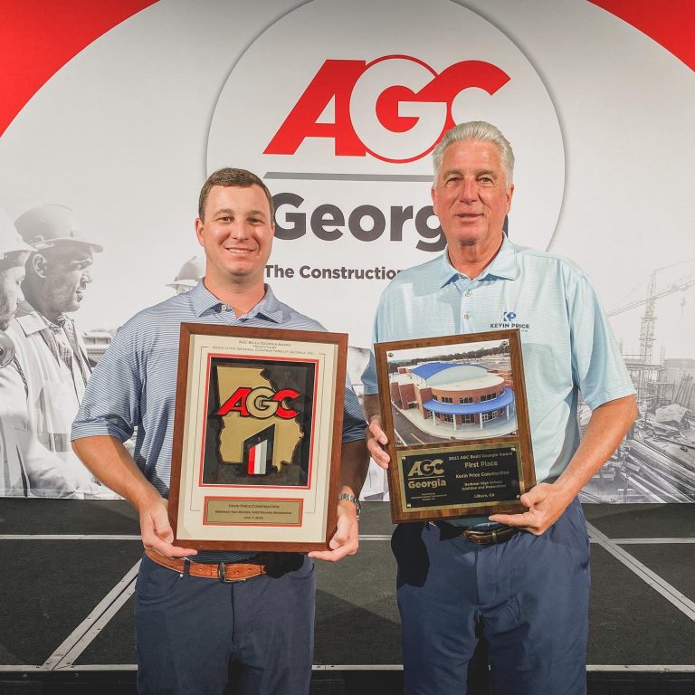 Two men stand on a stage holding AGC Georgia construction awards, with an AGC Georgia logo and construction-themed backdrop behind them.