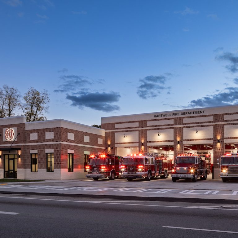 A modern brick fire station with four fire trucks parked in front; their lights are flashing. The building sign reads Hartwell Fire Department.