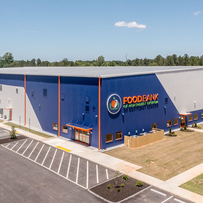 Large industrial warehouse building labeled Food Bank of Central & Eastern North Carolina with adjacent parking lot and surrounded by grass and trees under a clear sky.