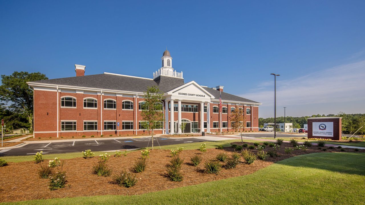 A two-story red brick building with white columns and a cupola, identified as Oconee County Schools, stands behind landscaped grounds and a sign.