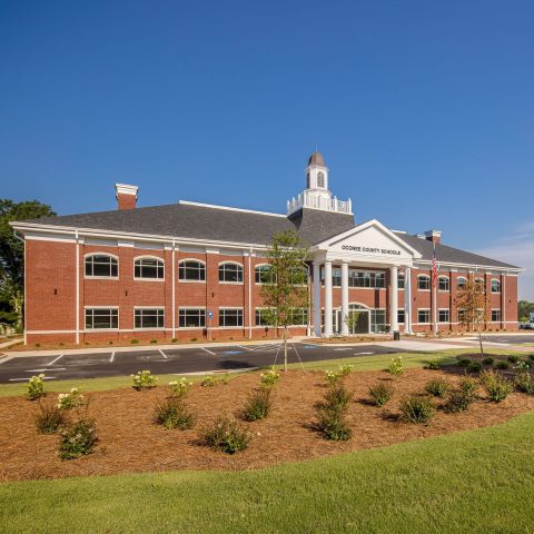 A two-story red brick building with white columns and a cupola, identified as Oconee County Schools, stands behind landscaped grounds and a sign.
