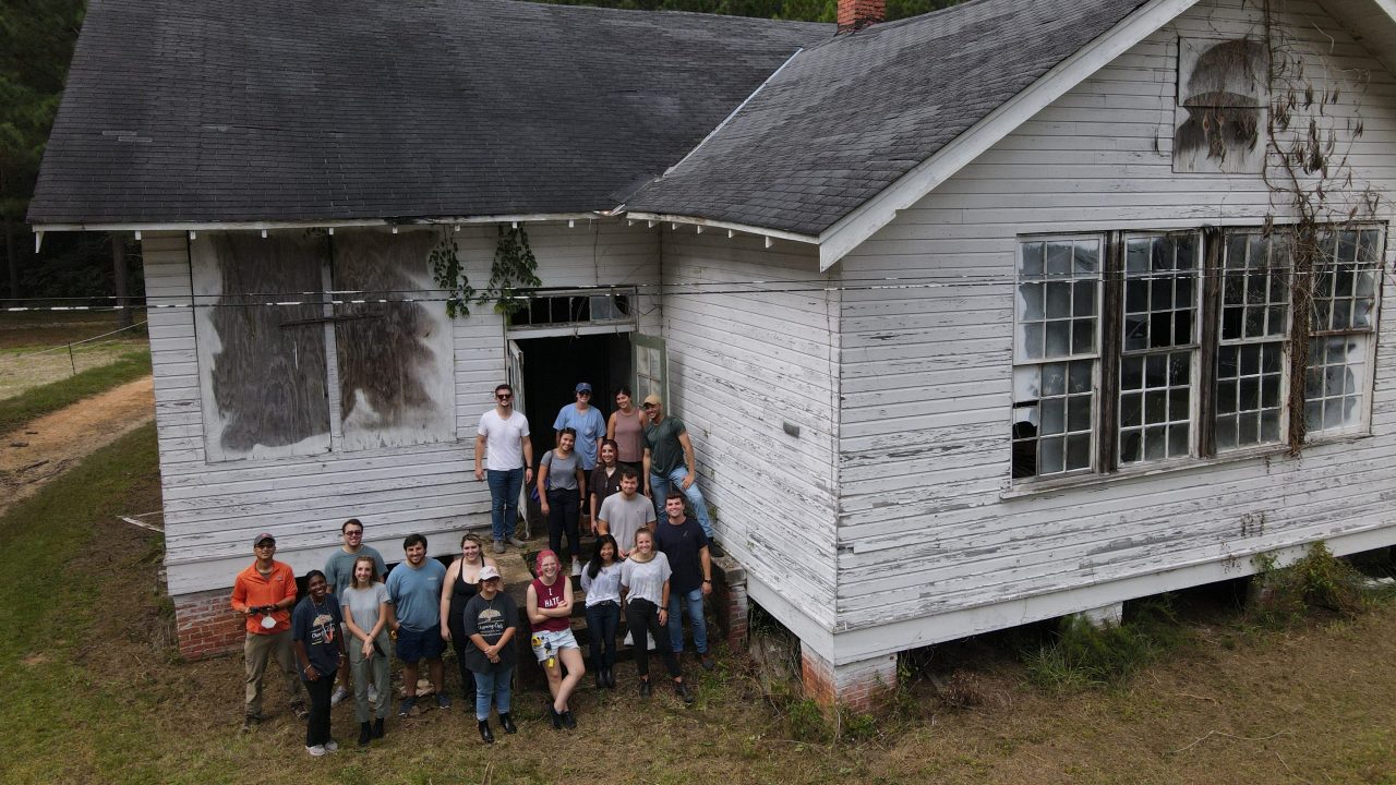 A group of people stands in front of a weathered, white wooden building with boarded-up windows and overgrown vegetation.