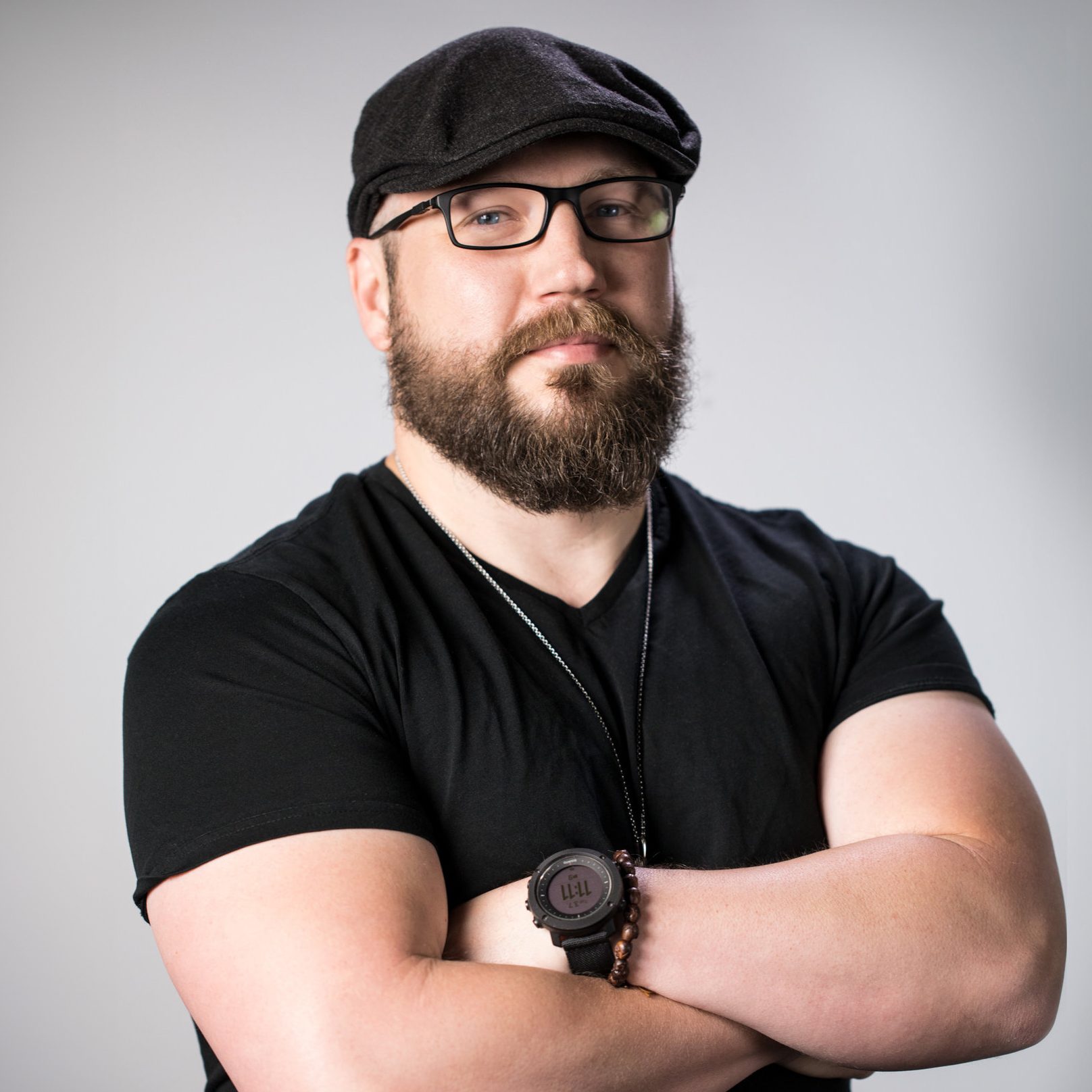 A man with a beard and glasses, wearing a black t-shirt, flat cap, and watch, stands with his arms crossed against a plain gray background.