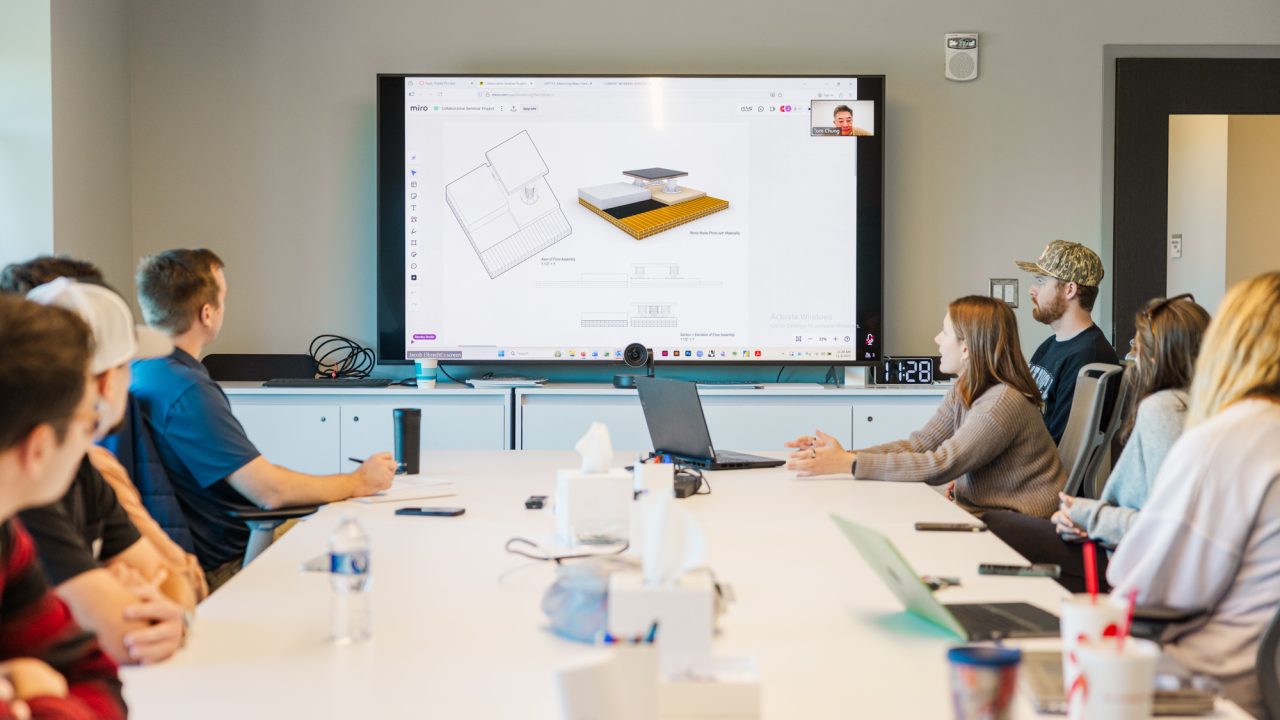 A group of people in a conference room watch a presentation on a large screen showing a 3D product design and a virtual meeting participant.