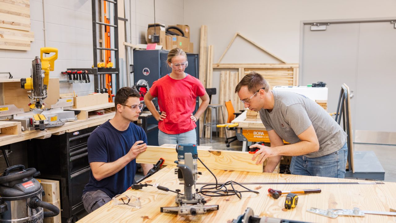 Three people work together on a woodworking project in a workshop, using tools and measuring a wooden plank on a large table.