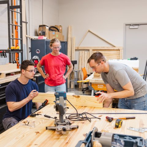 Three people work together on a woodworking project in a workshop, using tools and measuring a wooden plank on a large table.