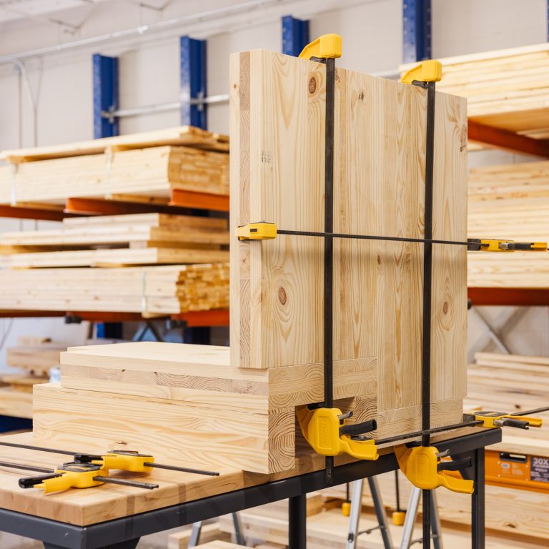 Several pieces of light-colored wood are clamped together on a worktable in a woodworking shop with stacked lumber in the background.