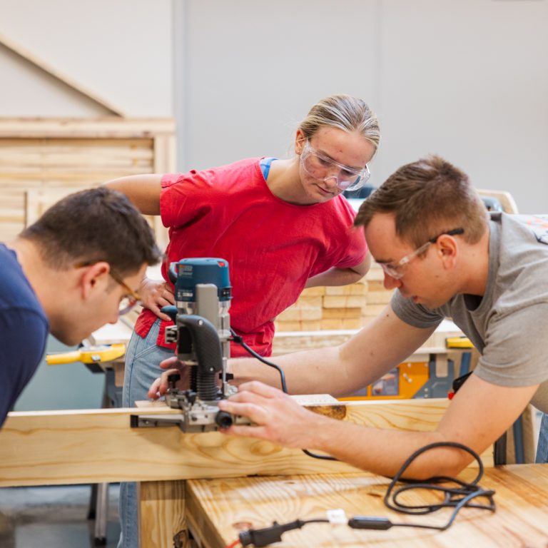 Three people wearing safety glasses work together on a woodworking project, using a power tool to cut a piece of wood in a workshop.