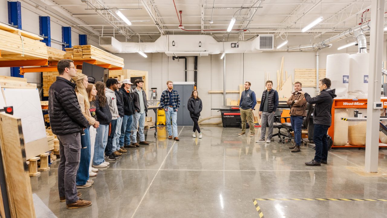 A group of people stand in a semi-circle inside a workshop with woodworking equipment, lumber, and tools visible in the background.