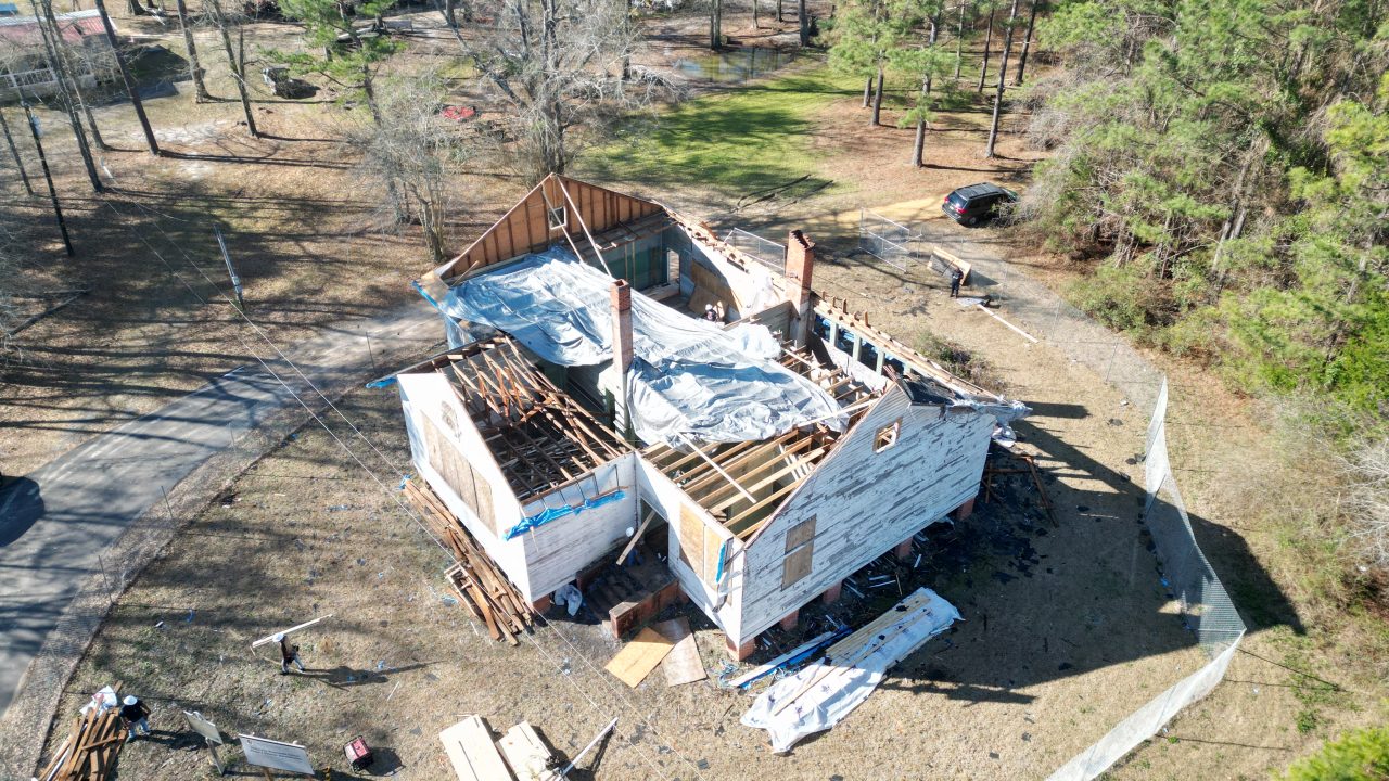 Aerial view of a partially demolished house with missing roof sections and debris scattered around the property, surrounded by trees and a grassy area.