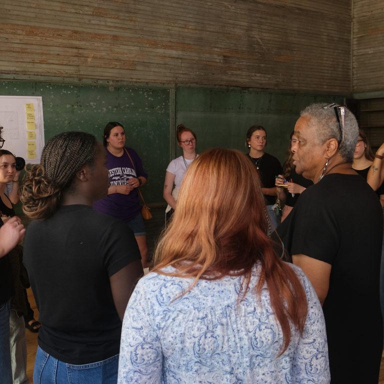 A group of people stand in a circle in a room with green chalkboards and wooden walls, engaging in discussion near a whiteboard with sticky notes.