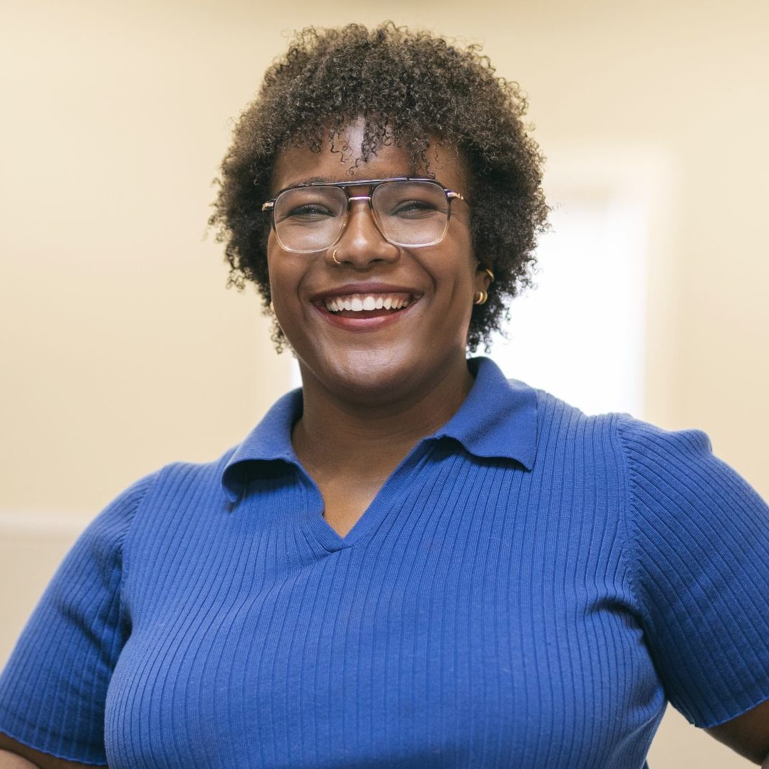 A person wearing glasses and a blue shirt is smiling and standing in a modern office setting.