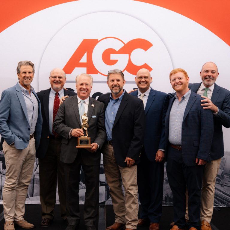 Seven men in business attire stand together smiling, with one man holding a trophy, in front of an AGC-branded backdrop.