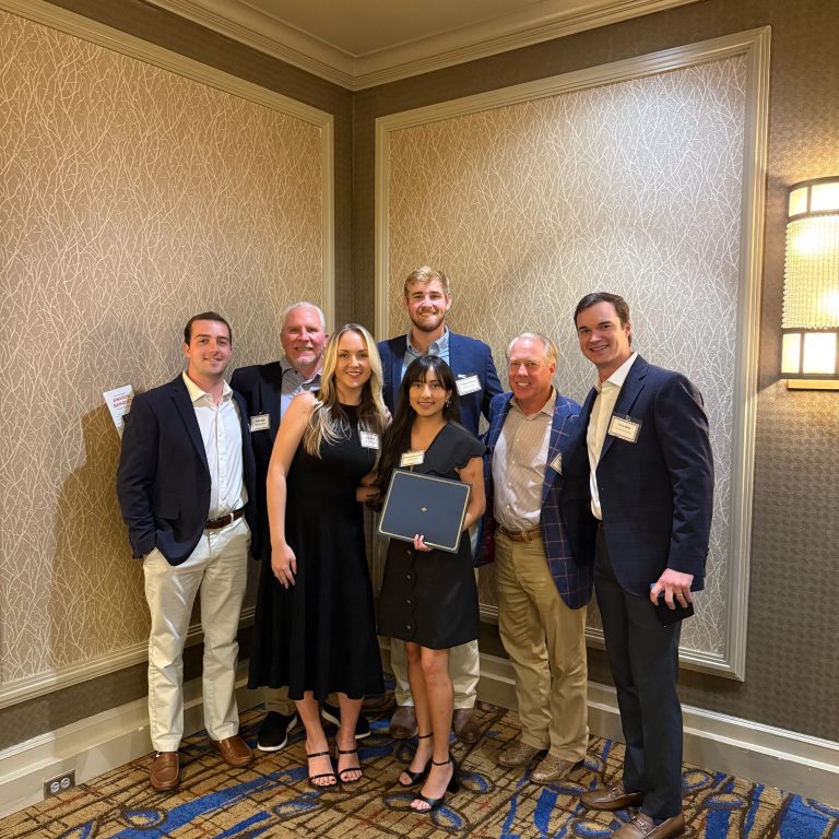 Six people posing indoors; one woman in the center holds a certificate. All are dressed in business or business casual attire, standing against a patterned wall and carpeted floor.