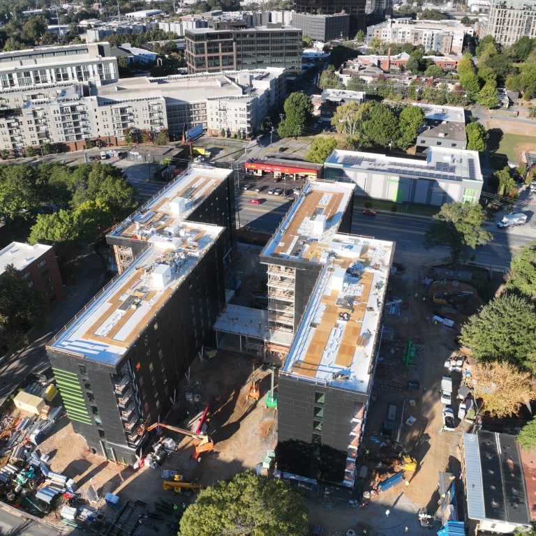 Aerial view of a mid-rise building under construction, surrounded by trees, equipment, and nearby residential and commercial structures.