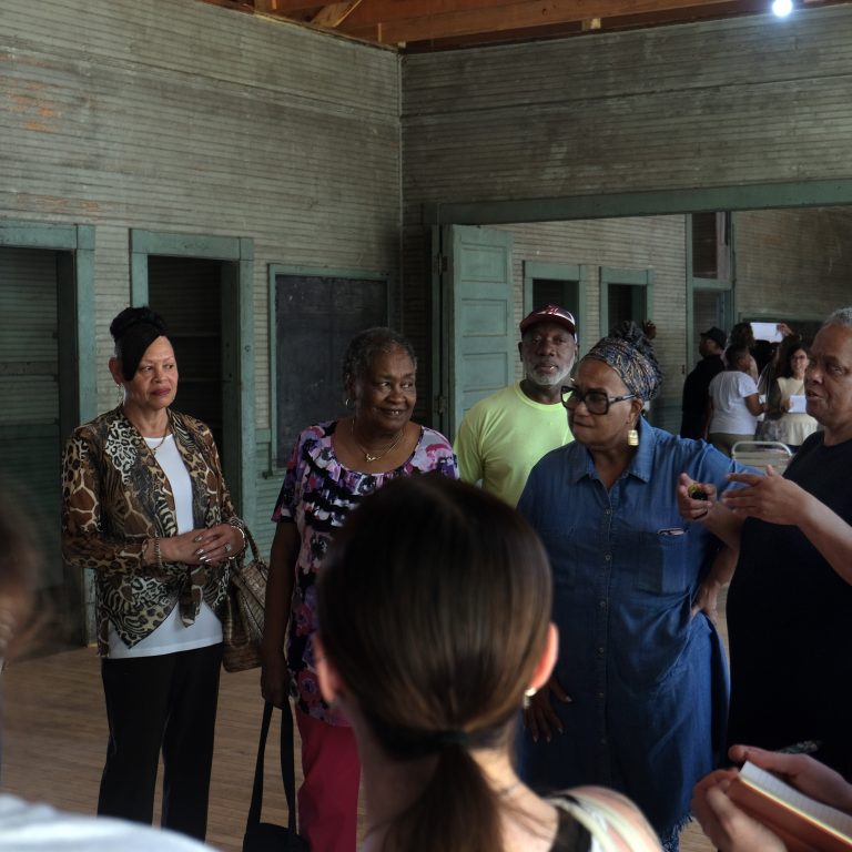A group of people stand and converse inside a rustic wooden building with green doors and exposed beams.