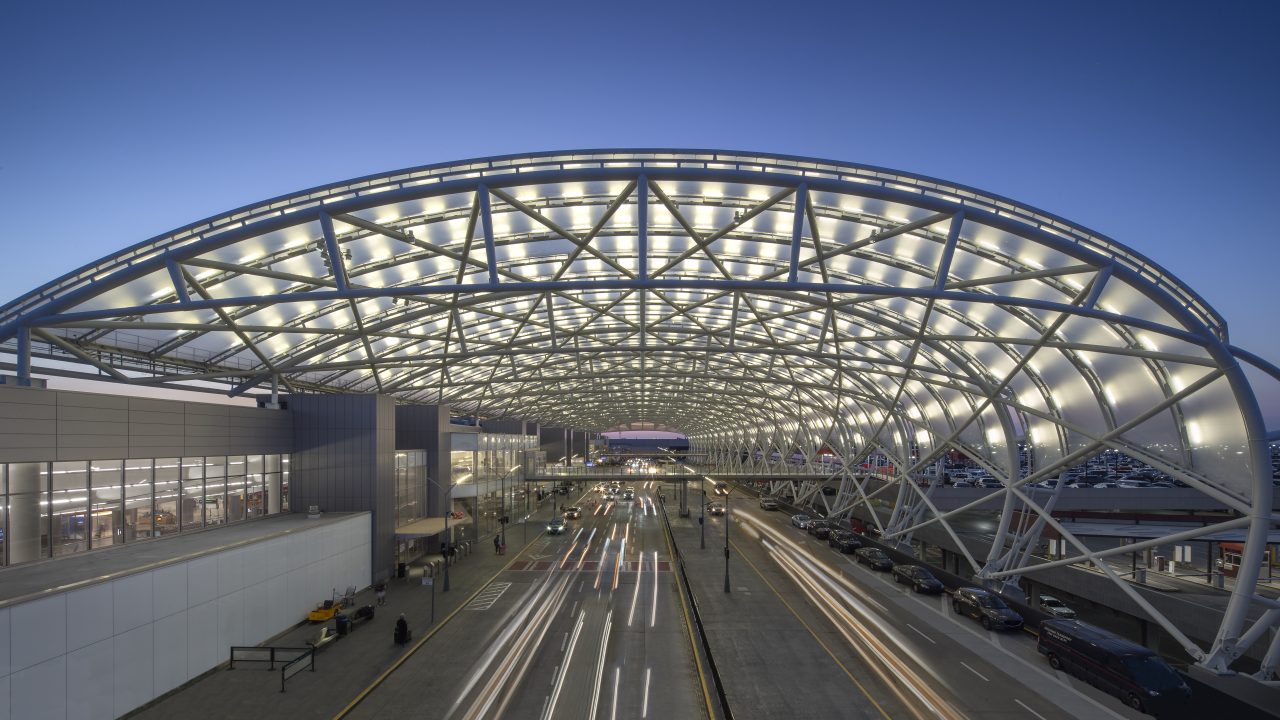 A modern airport terminal with an illuminated arched roof structure, cars and buses lined up at the entrance, and travelers moving along the sidewalk at dusk.
