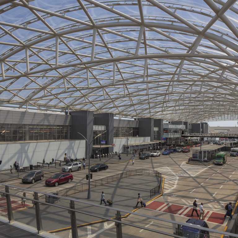 Travelers and vehicles outside a modern airport terminal with a large, curved glass and steel canopy overhead.