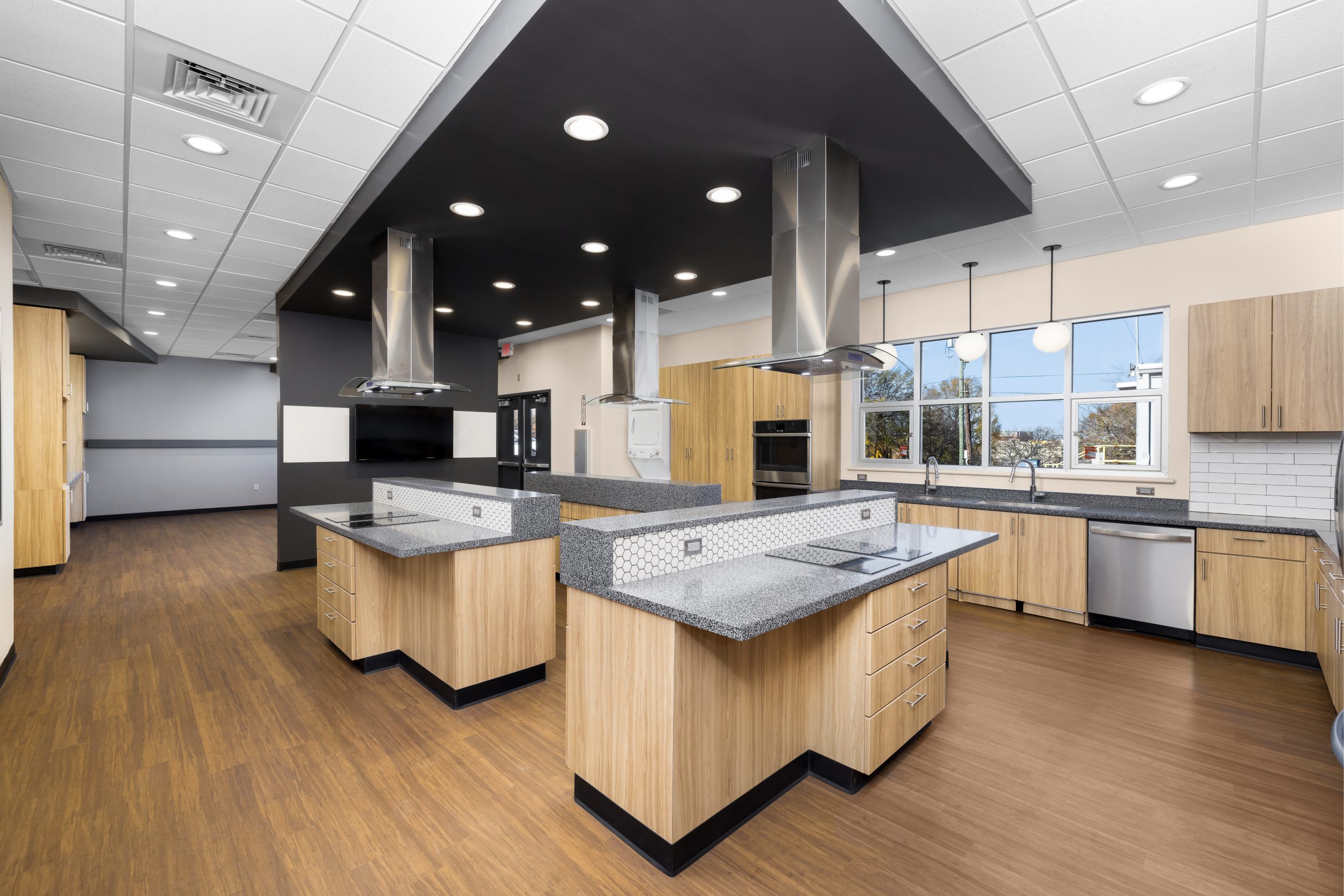 Modern commercial kitchen with wooden cabinets, multiple islands, stainless steel appliances, and large windows letting in natural light.