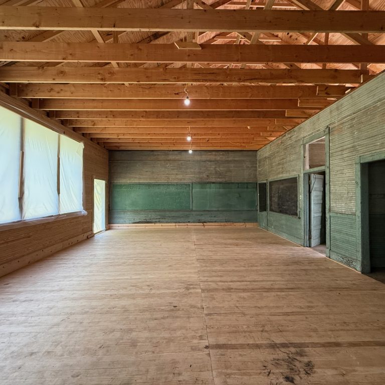 Large, empty room with exposed wooden beams, plywood floor, covered windows on the left, and green chalkboards on the back and right walls.