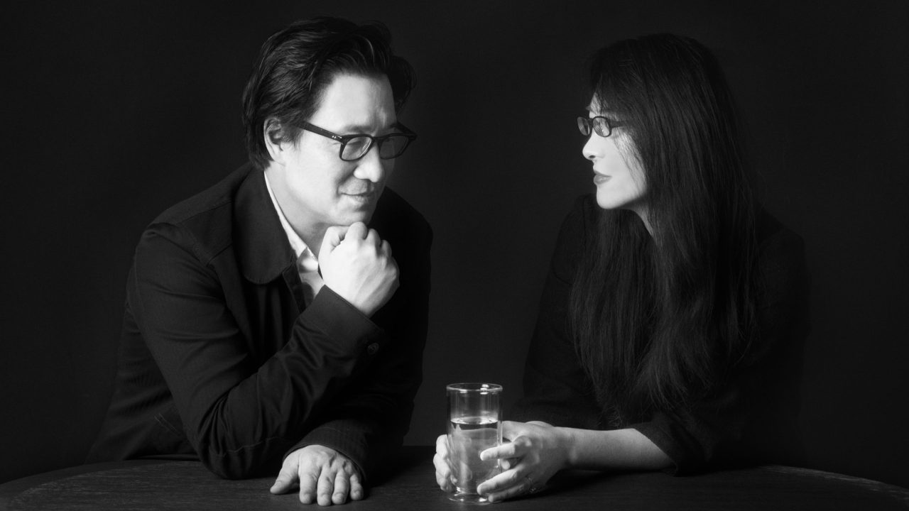A man and a woman sit at a round table against a dark background; the man rests his chin on his hand while the woman holds a glass of water and looks at him.