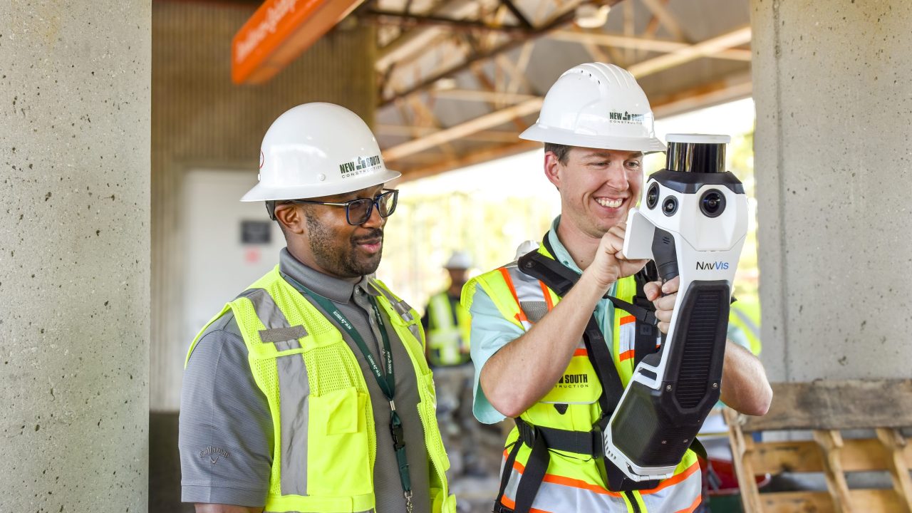 Two construction workers in safety vests and helmets use a handheld scanning device at a work site, with others and equipment visible in the background.