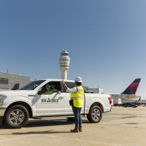 A construction worker in a safety vest stands by a white truck on an airport tarmac, with planes and a control tower visible in the background.