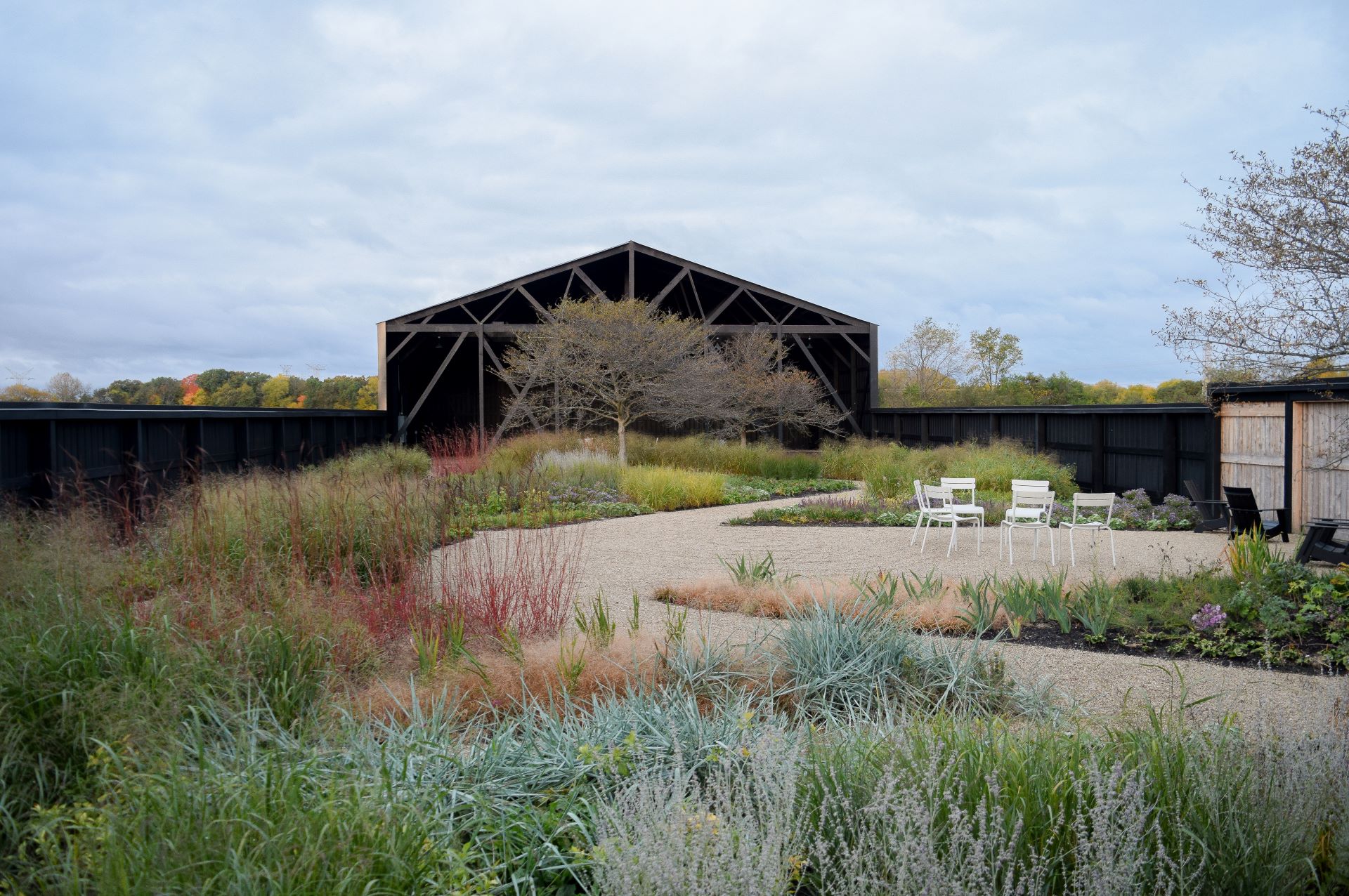A gravel path curves through a landscaped garden with grasses and plants, leading to a black modern barn structure. Three white chairs are arranged on the right side.
