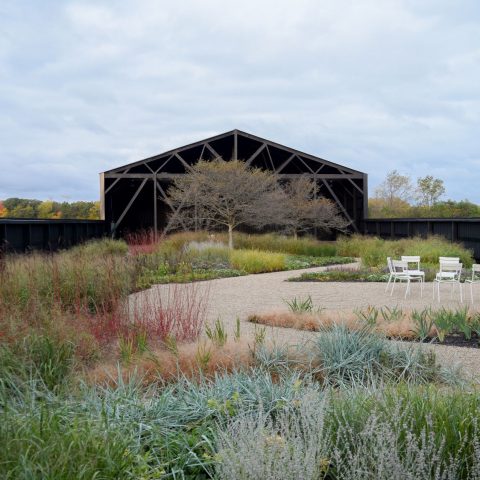 A gravel path curves through a landscaped garden with grasses and plants, leading to a black modern barn structure. Three white chairs are arranged on the right side.