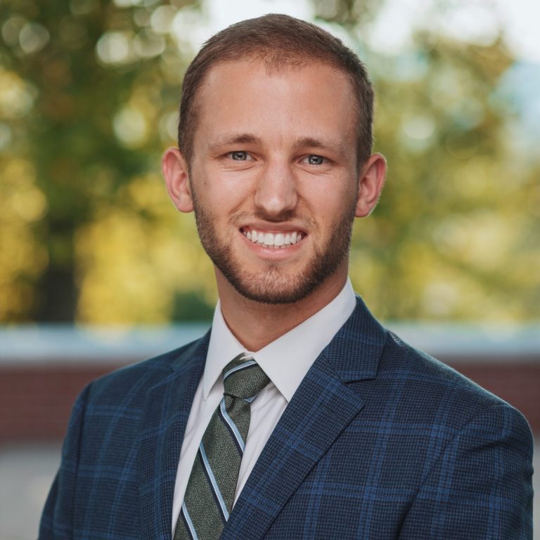 A man in a blue plaid suit, white shirt, and striped tie is standing outdoors, smiling at the camera, with trees in the background.